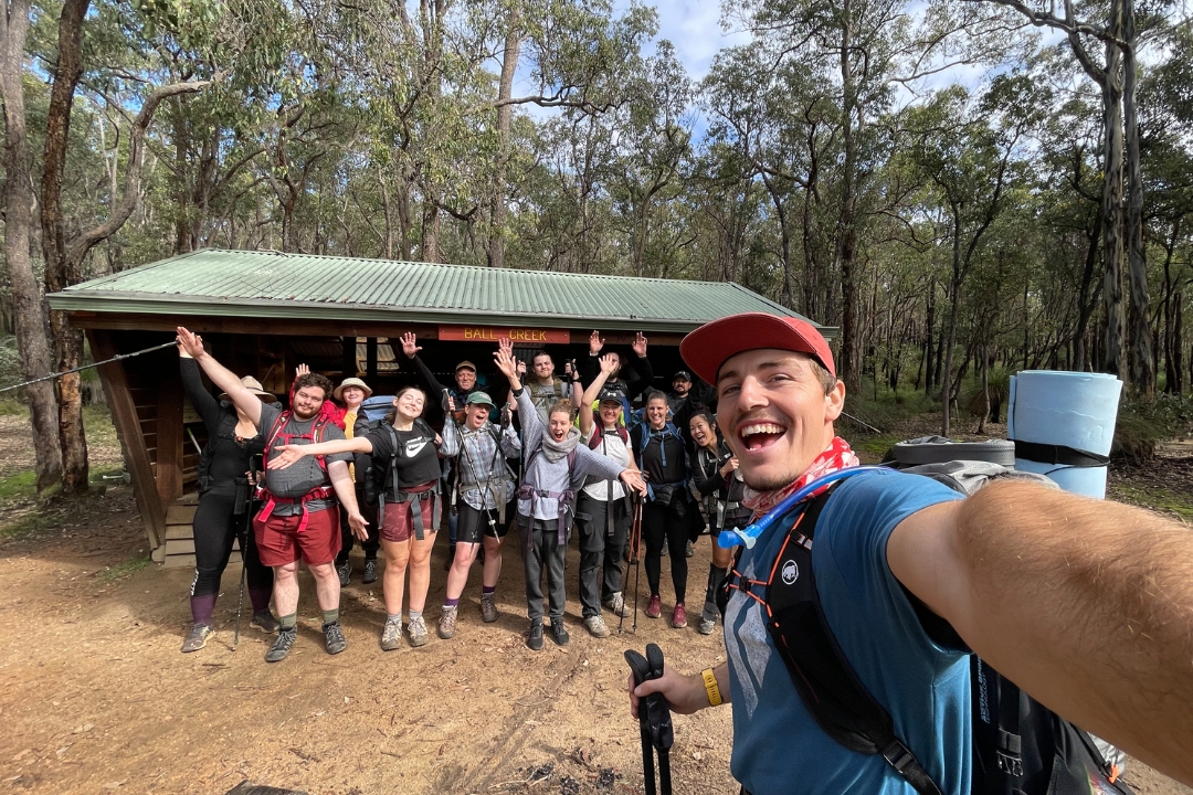 Group of people hiking, smiling at the camera in front of a shelter.