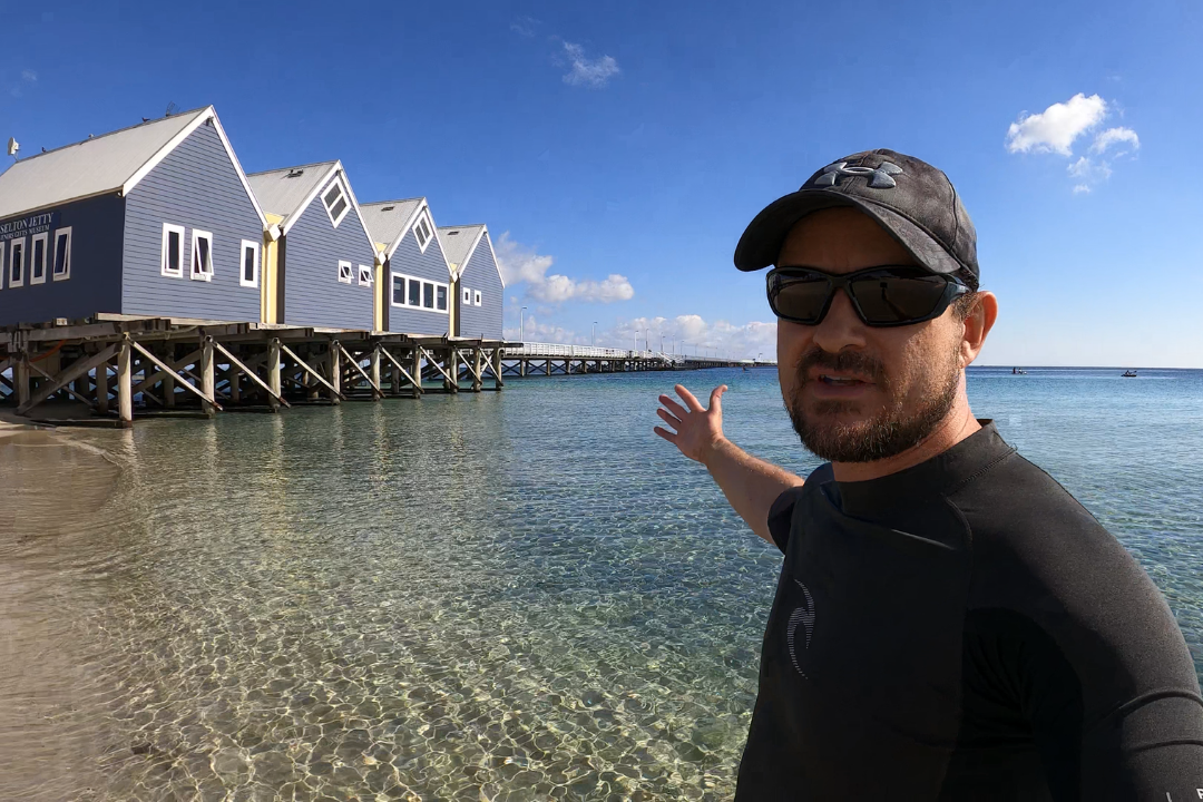 Man wearing baseball cap on the beach, pointing towards Busselton Jetty Interpretive Centre.